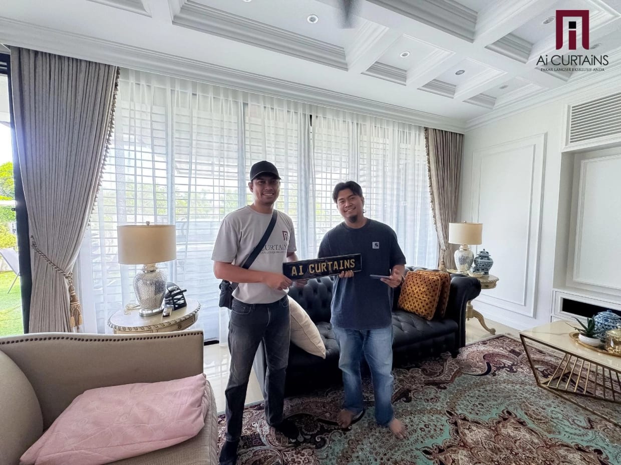 An Ai Curtains installer in company uniform standing with a customer in a finished living room, holding the company's plaque. Sheer curtains and heavy taupe drapes with tiebacks hang at the window behind them.