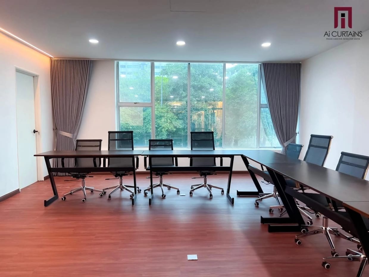 A long office meeting room with a full-height window, taupe floor-length drapes drawn to each side and a row of black office chairs down a light table.