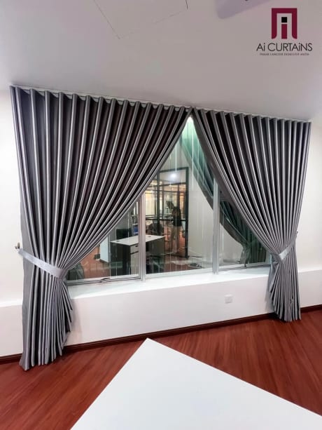 Grey ripplefold drapes with fabric tiebacks hanging beside a glazed office partition, over a timber floor.